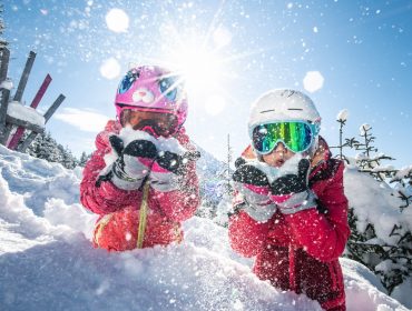 Dwoje dzieci w kaskach i goglach bawi się na śniegu, zdmuchując płatki śniegu, za którymi świeci słońce.