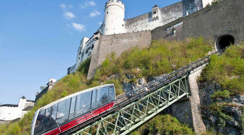 Czerwona kolejka linowa wspina się na strome wzgórze w Lato Salzburg w kierunku białej fortecy na szczycie kamiennego muru.