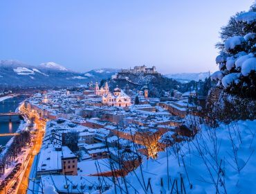 Pokryty śniegiem Salzburg o zmierzchu z rzeką, oświetlonymi ulicami i odległymi górami pod błękitnym niebem.