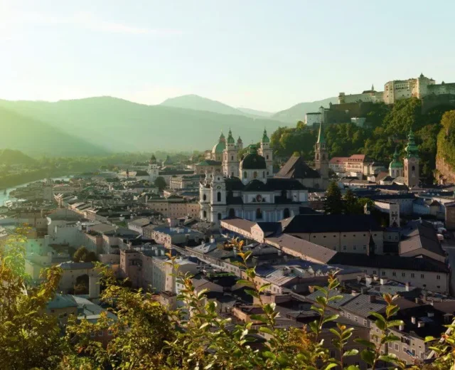 -salzburgerland-tourismus-1920×658.jpg Para obejmuje się, podziwiając malowniczy krajobraz Salzburga o zachodzie słońca, z górami w tle.