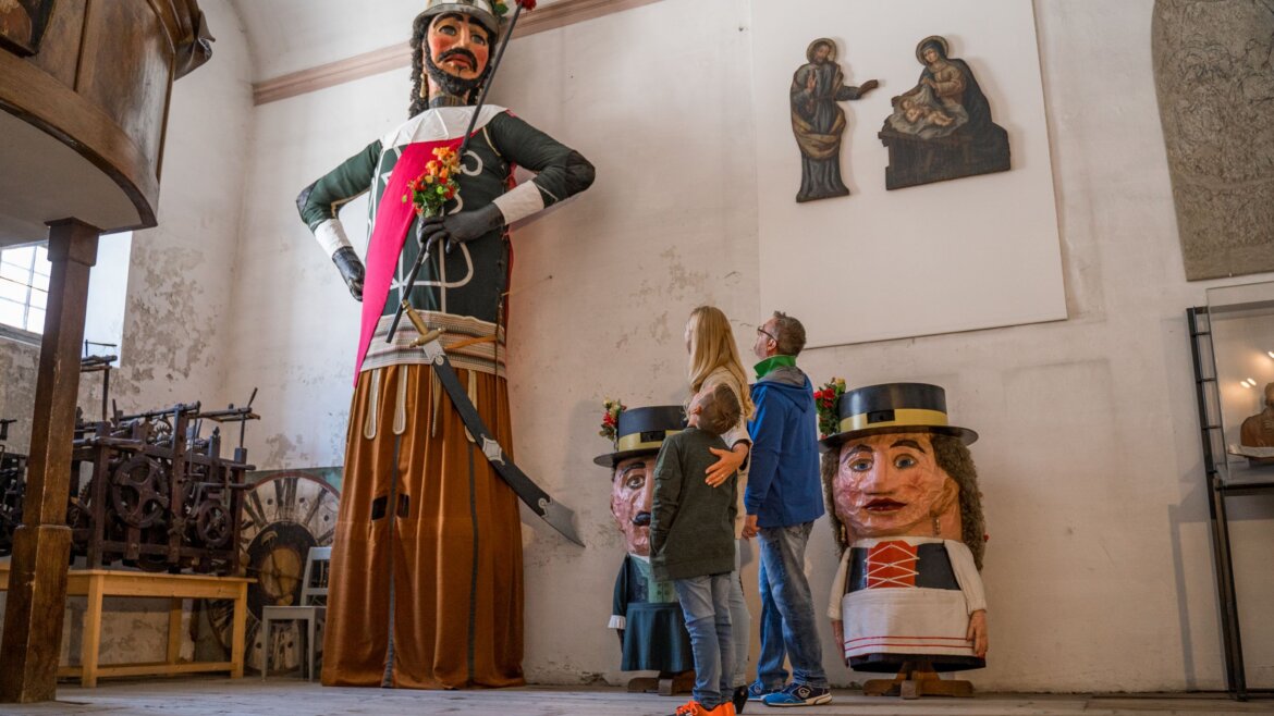 Dwie osoby podziwiają gigantyczne figury inspirowane zwyczaje ludowe Salzburski Lungau w białej sali muzeum.