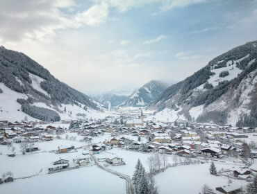 Pokryta śniegiem wioska leży w dolinie Raurisertal między zalesionymi górami pod częściowo zachmurzonym niebem.