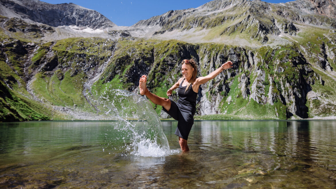 Kobieta pluskająca się stopą w czystym górskim jeziorze Wildkogel-Arena, w tle skaliste wzgórza.