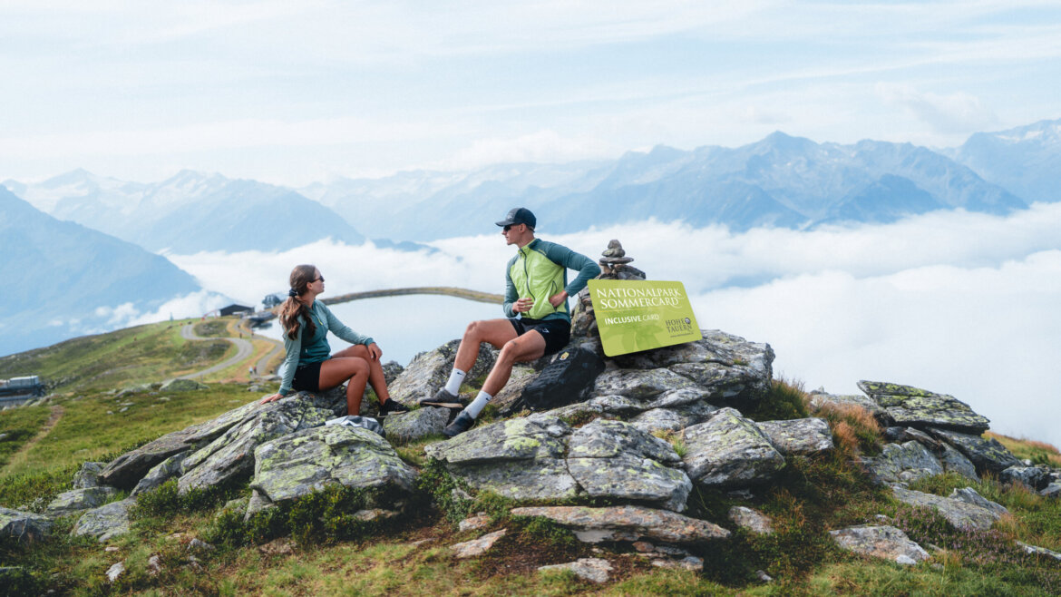 Dwóch turystów odpoczywa na skalistym terenie przy znaku Wildkogel-Arena, z górami i chmurami w tle.