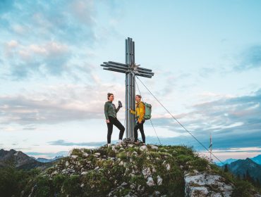 Dwóch wędrowców stoi przy dużym metalowym krzyżu na szczycie Obertauern pod błękitnym, zachmurzonym niebem.