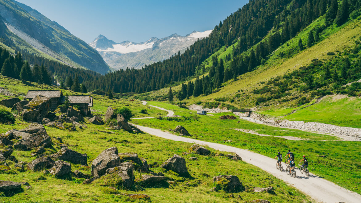Rowerzyści jadą krętą ścieżką przez zieloną górską dolinę Wildkogel-Arena z odległymi ośnieżonymi szczytami.