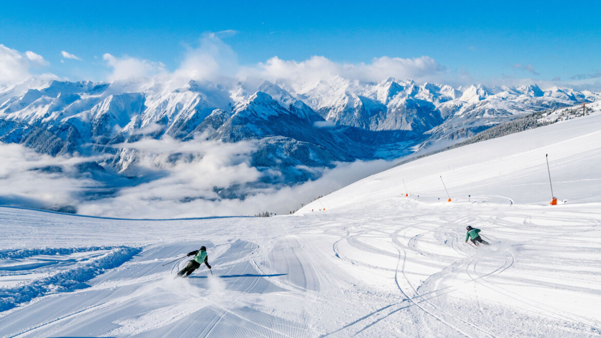 Dwóch narciarzy zjeżdża po ośnieżonym stoku w Wildkogel-Arena, otoczeni majestatycznymi szczytami i czystym, błękitnym niebem.