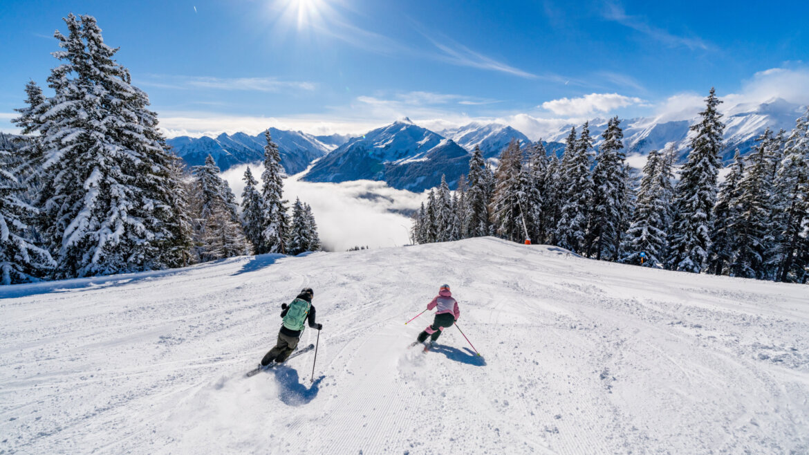 Dwie osoby zjeżdżają na nartach po ośnieżonych stokach Wildkogel-Arena, otoczone oświetlonymi słońcem szczytami i ośnieżonymi drzewami.
