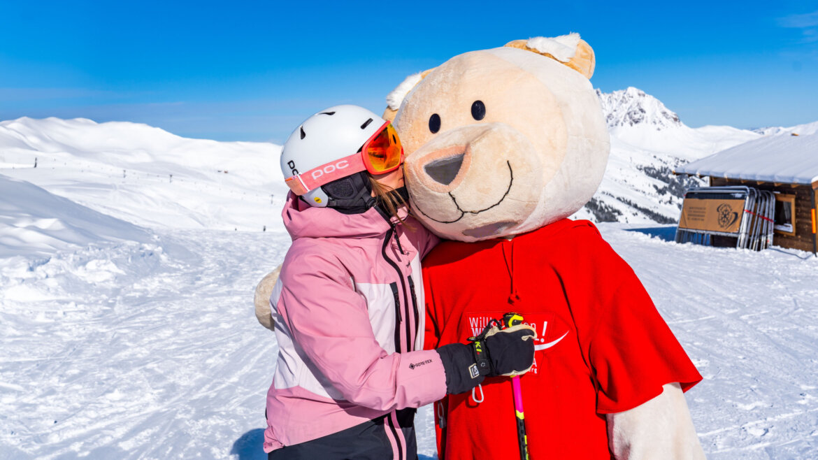 Narciarz w różowym stroju przytula osobę w kostiumie niedźwiedzia na Wildkogel-Arena pod czystym, błękitnym niebem.