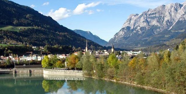 Bischofshofen Sommer Rzeka z zielonymi drzewami, budynkami miejskimi i wysokimi górami w tle pod błękitnym niebem.