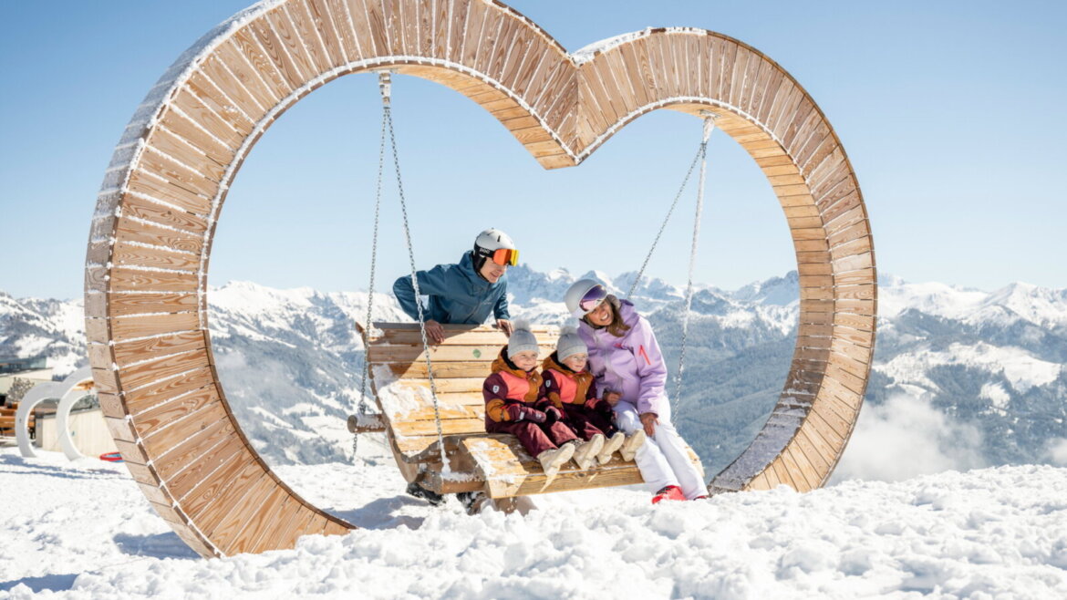 Słońce, góry, świeże powietrze - uwielbiają to dzieci i rodzice © TVB Grossarltal, Lorenz Masser Rodzina siedząca na huśtawce w kształcie serca, otoczona ośnieżonymi górami i błękitnym niebem. (Powiększony widok)
