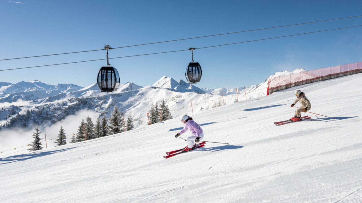 Na nartach zjazdowych na stokach w Grossarltal © TVB Grossarltal, Lorenz Masser Dwie osoby zjeżdżające na nartach pod wyciągami gondolowymi z ośnieżonymi górami i czystym błękitnym niebem w tle. (Powiększony widok)