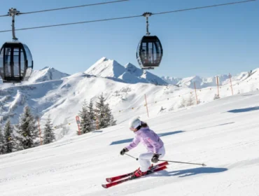 Dwie osoby zjeżdżające na nartach po ośnieżonym stoku górskim z kolejkami linowymi i ośnieżonymi szczytami w tle.