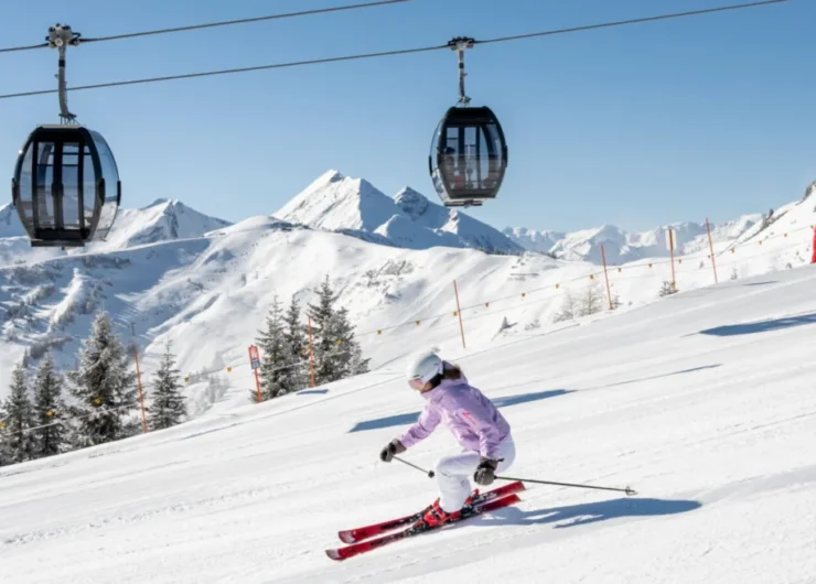 grossarltal-skifahren-gondelbahn–lorenzmasser-1920×658.jpg Dwie osoby zjeżdżające na nartach po ośnieżonym stoku górskim z kolejkami linowymi i ośnieżonymi szczytami w tle.