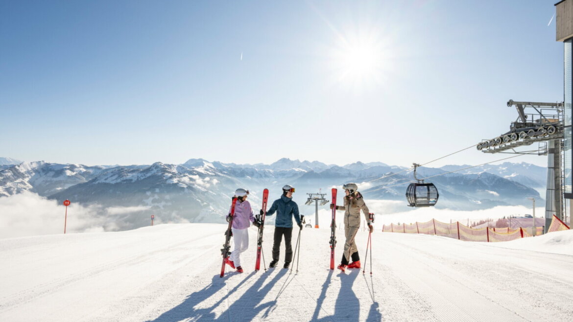 Wysoko nad doliną Grossarltal śnieg spotyka się ze słońcem © TVB Grossarltal, Lorenz Massert Czterech narciarzy stoi na ośnieżonym stoku z wyciągami narciarskimi i górskimi szczytami w tle w jasnym słońcu. (Powiększony widok)