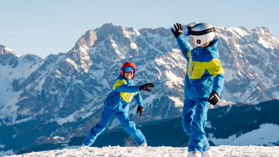 Region Hochkönig posiada atrakcjyną ofertę zimową dla dzieci © Hochkönig Tourismus Miriam Lottes Dwoje dzieci w niebieskich kombinezonach rzuca się śnieżkami na ośnieżonej górze Zima w Hochkönig ze skalistymi szczytami za plecami. (Powiększony widok)