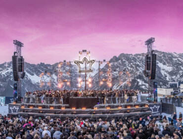 Tłum przed sceną festiwalu muzycznego Zima w Hochkönig z dużym logo w kształcie korony, na tle ośnieżonych gór o zachodzie słońca.