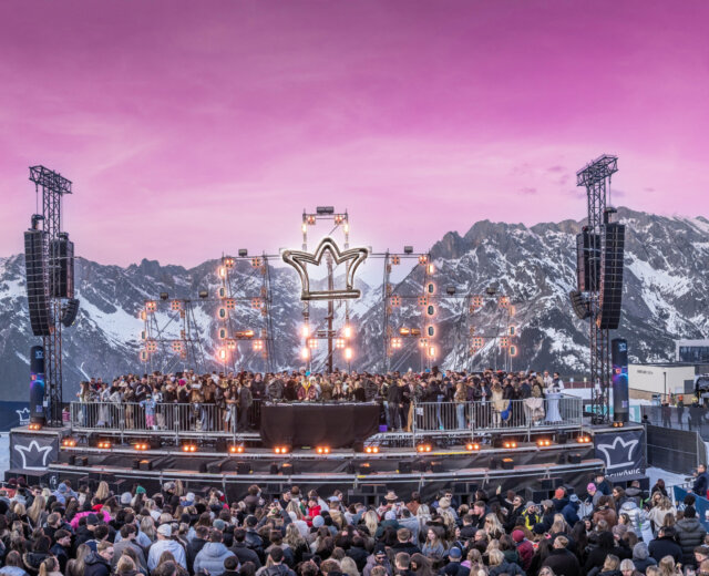 Tłum przed sceną festiwalu muzycznego Zima w Hochkönig z dużym logo w kształcie korony, na tle ośnieżonych gór o zachodzie słońca.