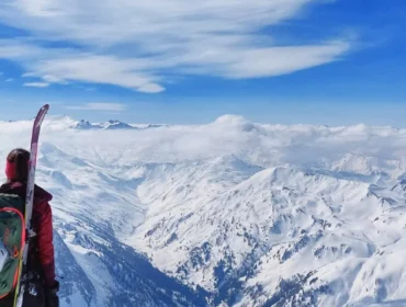 Osoba z nartami i plecakiem spogląda na ośnieżone szczyty Salzburskiego Lungau pod jasnoniebieskim niebem z chmurami.