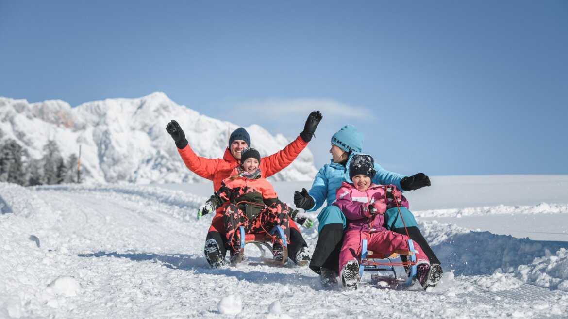 Sanki z rodziną w Hochönig © Hochkönig Tourismus Czteroosobowa rodzina cieszy się Zima w Hochkönig, zjeżdżając na sankach po zaśnieżonych stokach górskich pod czystym, błękitnym niebem. (Powiększony widok)