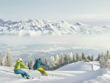 Dwie osoby zjeżdżające na nartach w Zima w Schladming-Dachstein, otoczone ośnieżonymi szczytami i zalesionymi stokami.