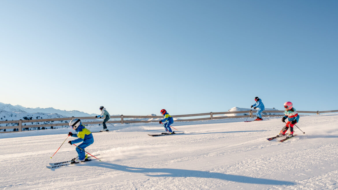 Narty z rodziną w regionie Hochkönig © Hochkönig Tourismus Miriam Lottes Czworo dzieci zjeżdżających na nartach pod czystym, błękitnym niebem - Zima w Hochkönig z górami w tle. (Powiększony widok)