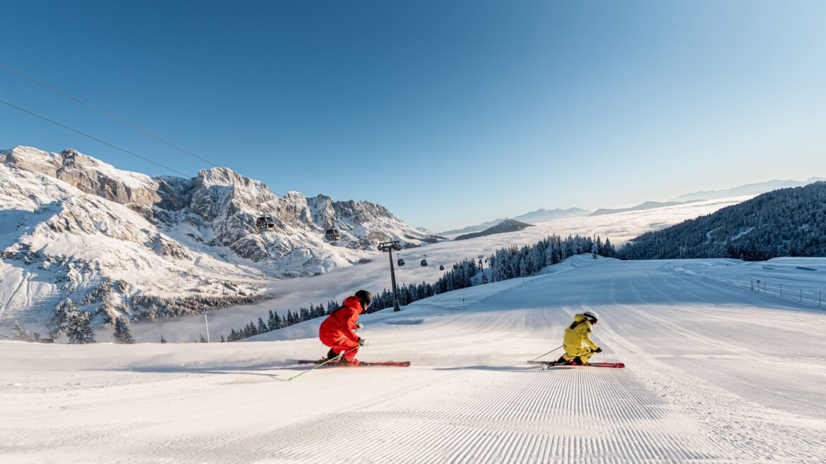 Szusowuj jak król w Mühlbach © Hochkönig Tourismus Dwóch narciarzy w jasnych strojach zjeżdża w dół podczas Zima w Hochkönig przy czystym niebie i malowniczych widokach. (Powiększony widok)