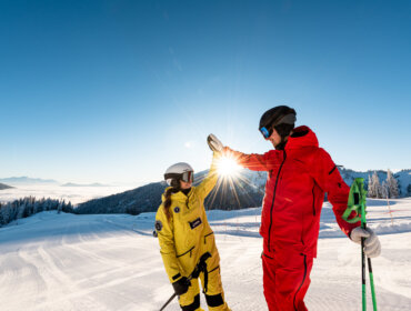 Dwóch narciarzy w jasnych kombinezonach przybija piątkę na słonecznym stoku Zima w Hochkönig pod czystym, błękitnym niebem.