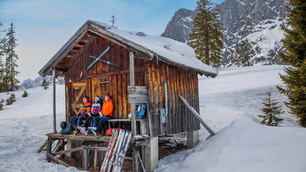 Przerwa na wypoczynek podczas wyprawy skitourowej w regionie Hochkönig © Hochkönig Tourismus, Johannes Felsch Dwie osoby w sprzęcie narciarskim relaksują się na zaśnieżonej werandzie domku podczas Zima w Hochkönig, narty i góry za nimi. (Powiększony widok)