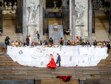 festspiele_jedermann-salzburg-2015_tsg-e1497884978526 Wielkie Sto Lat Festiwalu Salzburskiego z aktorami, długim stołem i tancerką w czerwieni z przodu.