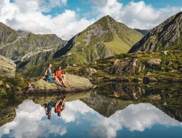schladming-dachstein-gartner_mathaus_-8031-1170×658 Dwie osoby relaksują się nad górskim jeziorem w Lato w Schladming-Dachstein, szczyty i chmury odbijają się w wodzie.