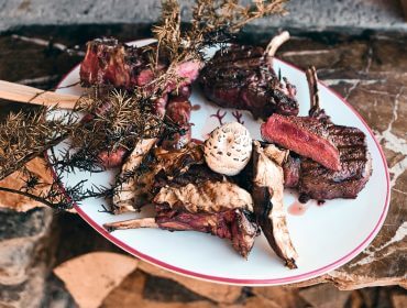 © Armin Walcher Talerz grillowanych kotletów z galaretką z grzybami, przyozdobiony ziołami, ustawiony na rustykalnej kamiennej powierzchni.