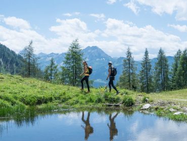 Dwie osoby wędrujące obok małego stawu w zielonym górskim krajobrazie z drzewami i błękitnym niebem.