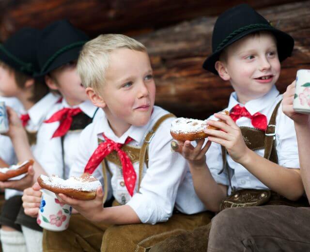 Bauernherbst – Kinder / Kulinarik Dzieci w tradycyjnych austriackich strojach delektują się wypiekami Hity wiejskiego stołu, siedząc w rzędzie na zewnątrz.