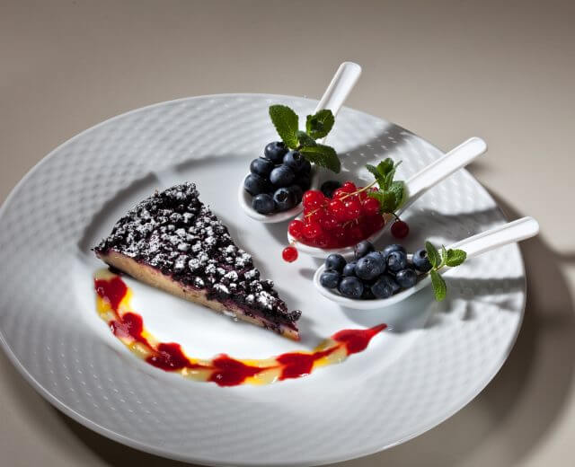 Gerührter Heidebeerkuchen Kawałek ciasta ucieranego z jagodami, z cukrem pudrem, jagodami i miętą na białym talerzu z sosem.