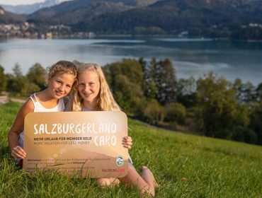 Dwie dziewczyny siedzą na trawiastym wzgórzu nad jeziorem, uśmiechają się i trzymają kartę SalzburgerLand.