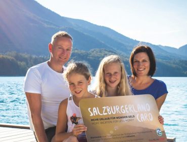 rs2995_2018-09-21-sl-card-sujet-by-michael-groessinger-img_5217-bearbeitet-lpr-1170×658 Uśmiechnięta rodzina pozuje nad jeziorem z górami, trzymając znak FAQ odnośnie SalzburgerLand Card.