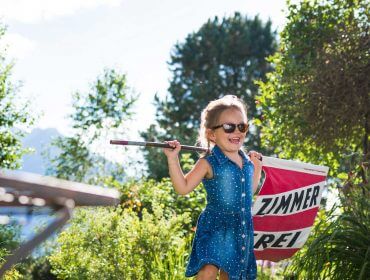 alpine_gastgeber_david_innerhofer Uśmiechnięta dziewczyna w okularach przeciwsłonecznych niesie znak Zimmer Frei dla Stowarzyszenia prywatnych kwaterodawców w słonecznym ogrodzie.