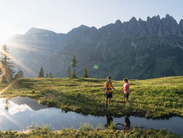 Dwóch wędrowców nad małym stawem w Lato w Ski amadé, z flarą słońca, trawiastym polem i surowymi górami za plecami.