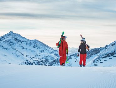 first-track_obertauern-1170×658-1 Dwóch narciarzy w czerwonych strojach spaceruje po zaśnieżonej górze z nartami w Narty w Obertauern, w otoczeniu szczytów.