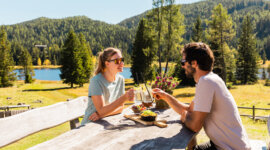 Para cieszy się atrakcjami kulinarnymi Salzburski Lungau, popijając drinki przy stoliku nad jeziorem pośród drzew.
