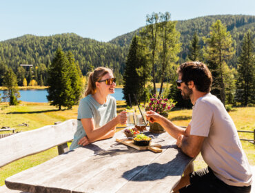Para cieszy się atrakcjami kulinarnymi Salzburski Lungau, popijając drinki przy stoliku nad jeziorem pośród drzew.