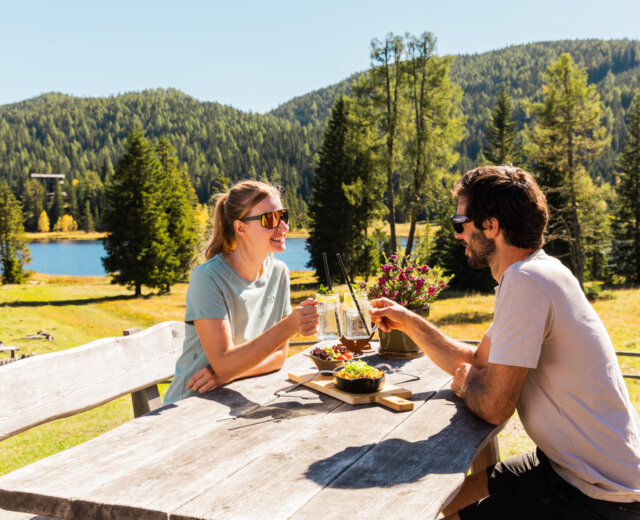 Para cieszy się atrakcjami kulinarnymi Salzburski Lungau, popijając drinki przy stoliku nad jeziorem pośród drzew.