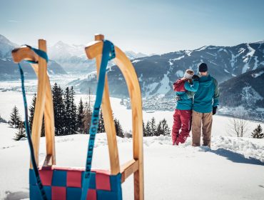 Para przytula się w zimowych ubraniach, z widokiem na ośnieżone góry z sankami w Zell am See-Kaprun.