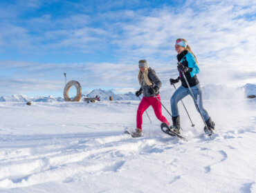 Dwie kobiety na rakietach śnieżnych w górach, cieszące się zimowymi wędrówkami w Wildkogel-Arena pod słonecznym niebem.