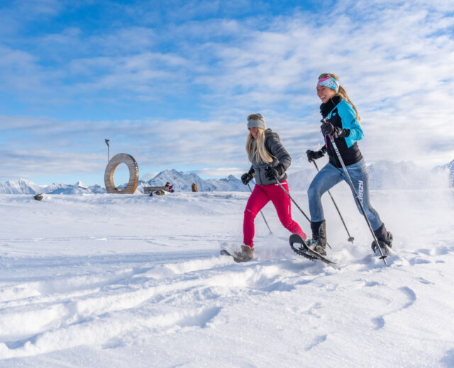 Dwie kobiety na rakietach śnieżnych w górach, cieszące się zimowymi wędrówkami w Wildkogel-Arena pod słonecznym niebem.