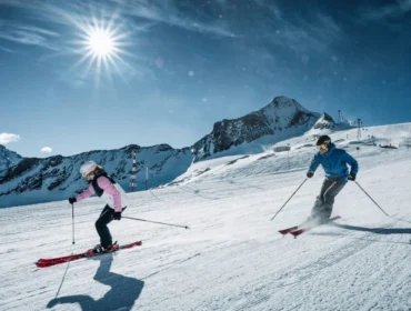 Dwie osoby zjeżdżające na nartach z ośnieżonej góry, cieszące się imprezą w Zell am See-Kaprun w jasnym słońcu.