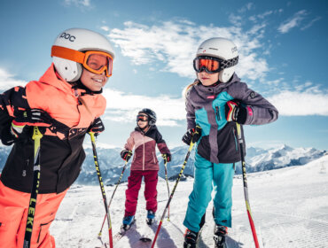 Trójka dzieci w sprzęcie narciarskim jeździ na nartach i uśmiecha się na zaśnieżonej górze podczas ferie zimowe z rodziną w Zell am See-Kaprun.