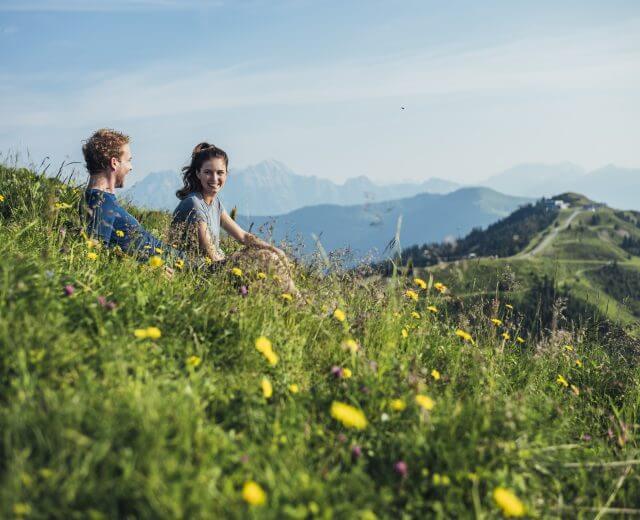 wanderpause-auf-der-schmittenhohe—hiking-break-on-schmittenhohe-c-zell-am-see-kaprun-tourismus Dwie osoby siedzą na trawiastej łące z żółtymi kwiatami, z widokiem na góry pod czystym niebem.