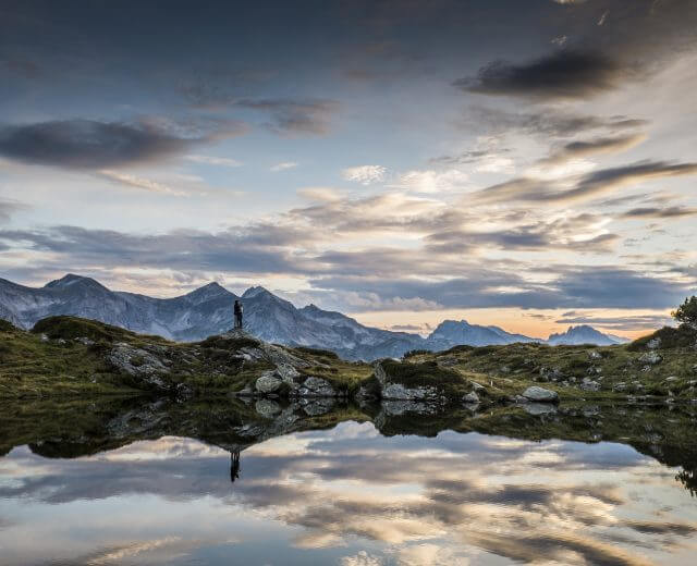 Osoba stoi nad jeziorem z górami i dramatycznym, zachmurzonym niebem odbijającym się w wodzie o zachodzie słońca.